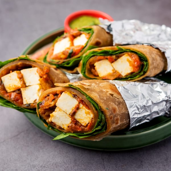 Indian paneer kathi rolls, some wrapped in foil, on a green plate with a side of green chutney.